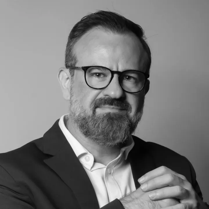 Retrato em preto e branco de Mário Trentim, de óculos e barba, com paletó escuro e as mãos unidas à frente